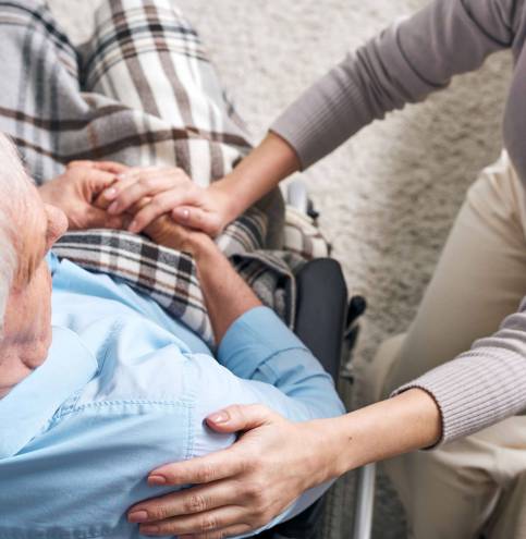 Young supportive female caregiver sitting by senior man in wheelchair and keeping her hand on his shoulder while comforting him